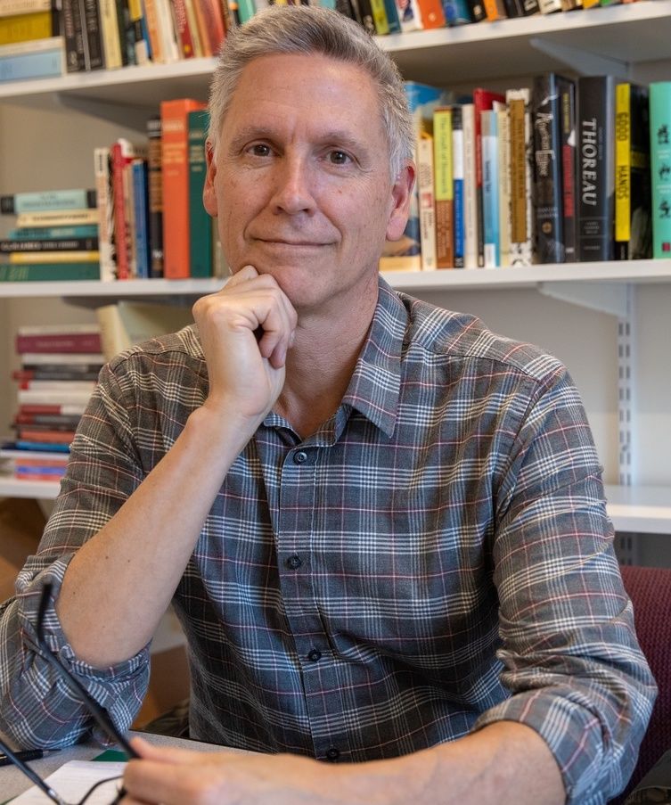 Photo of person wearing plaid shirt against bookcase looking into camera with one hand to the chin